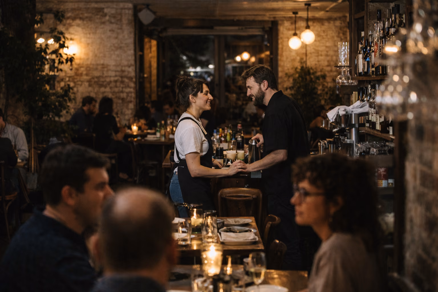 Bartender and waitress smiling and holding hands in a warmly lit, busy restaurant bar.