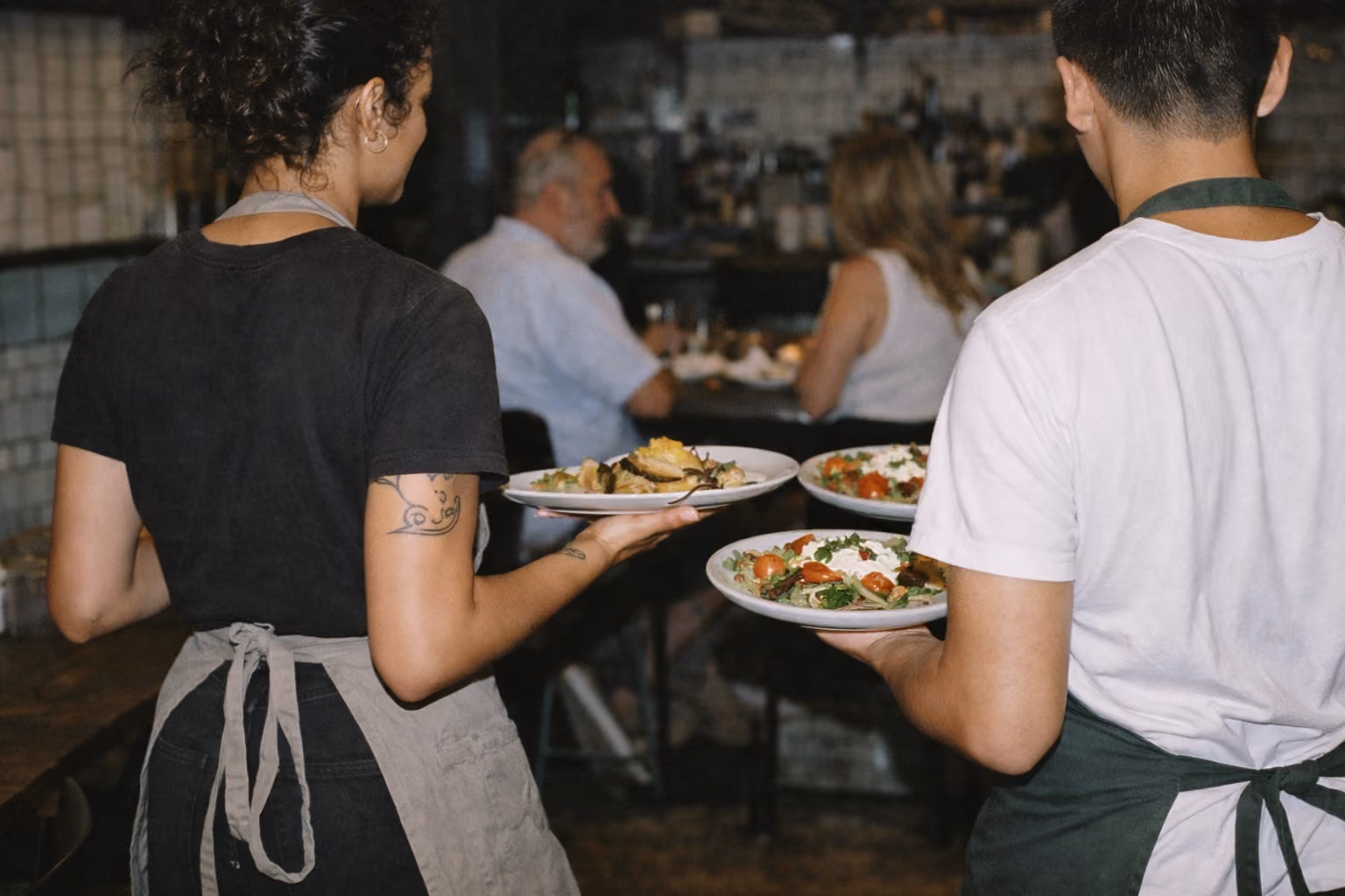 Two servers carrying plates of food in a busy restaurant with seated diners in the background.
