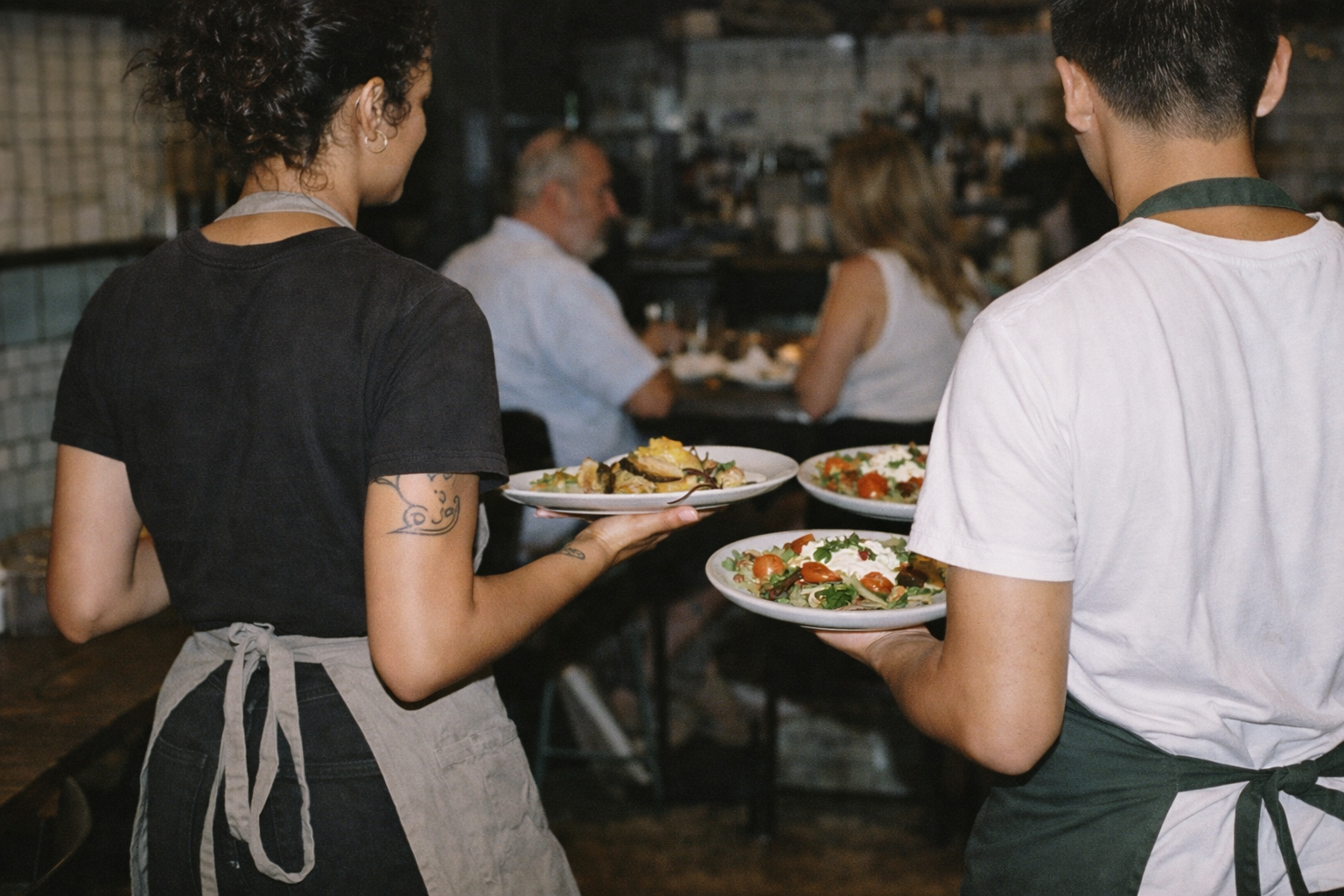 Two servers carrying plates of food in a busy restaurant with seated diners in the background.