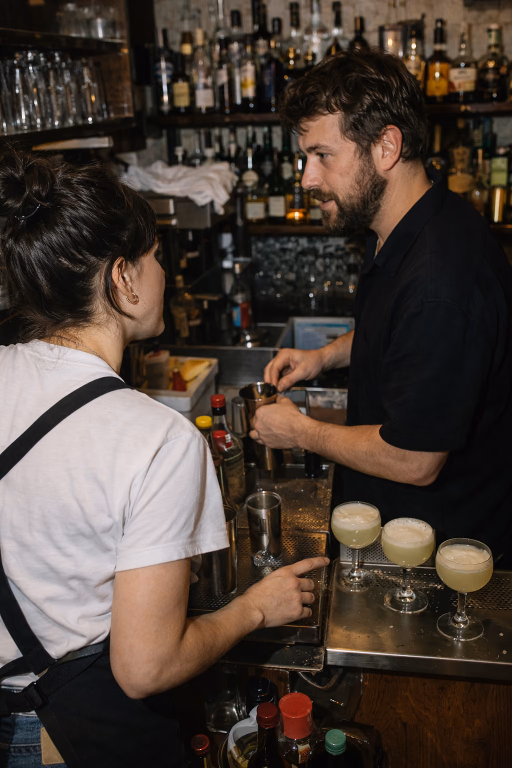 Female bartender talking to male bartender who is preparing cocktails behind the bar.