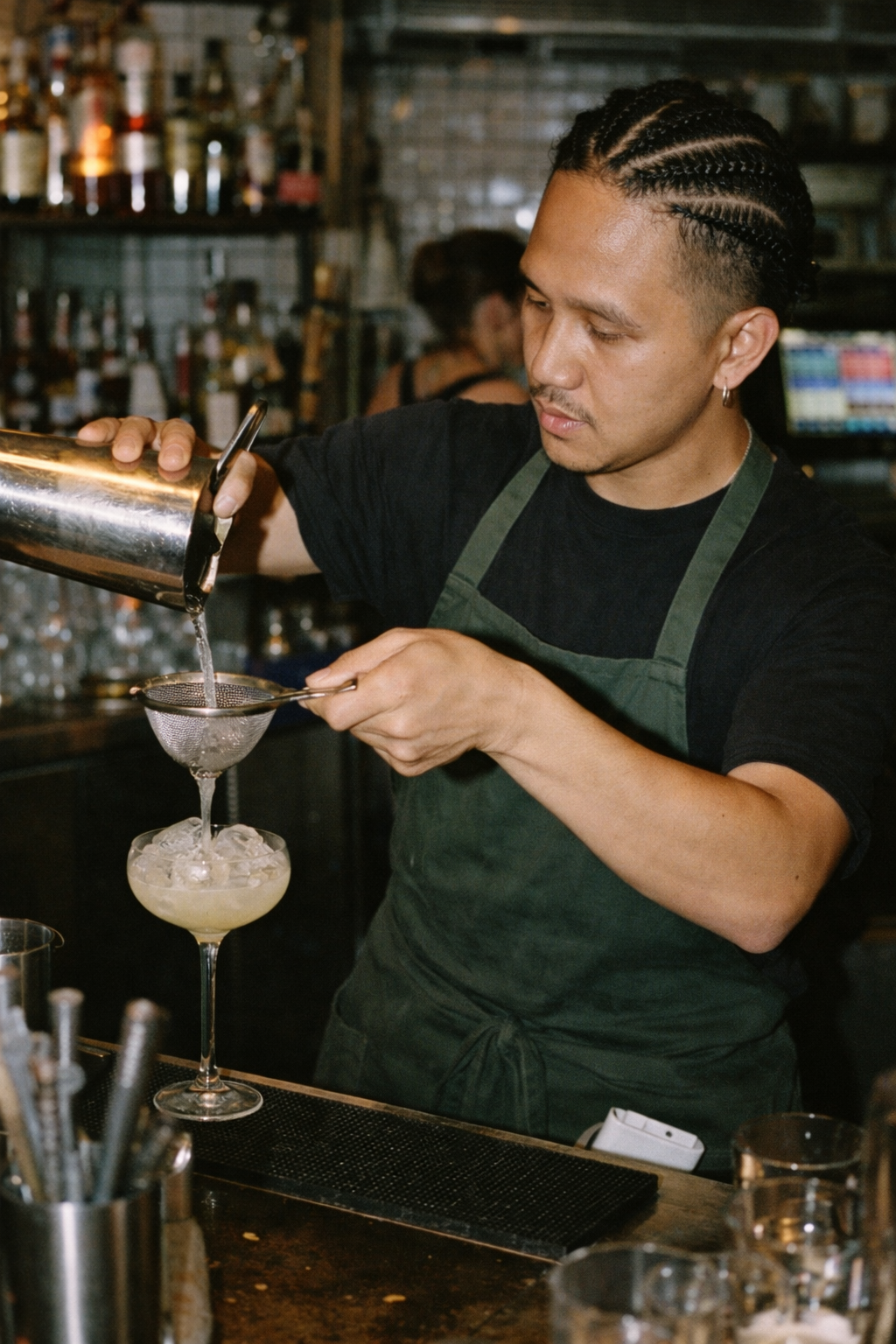 Bartender with braided hair pours cocktail through a strainer into a glass with ice at a bar.