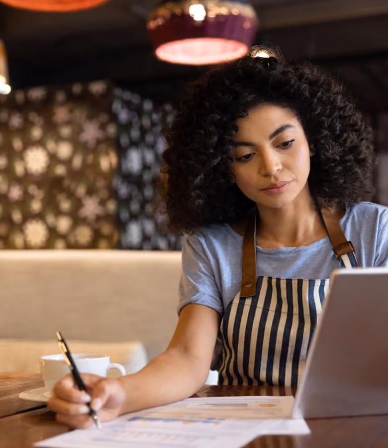 Woman in striped apron working on papers and tablet at a table with a cup of coffee.