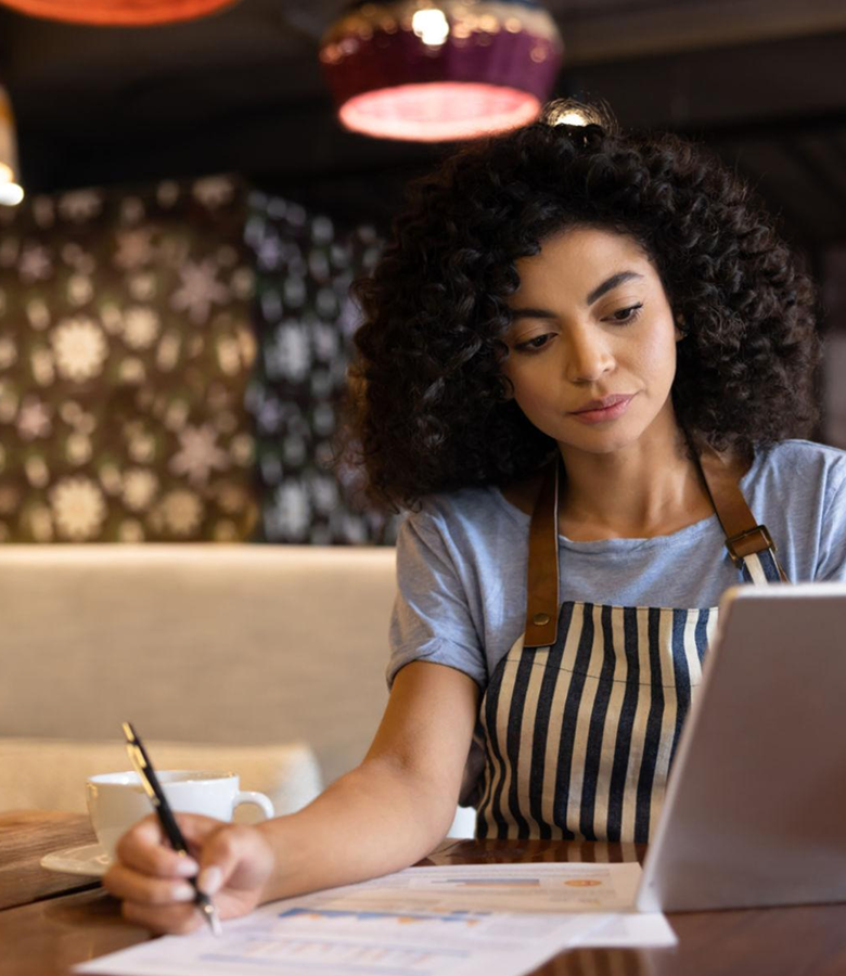 Woman in striped apron working on papers and tablet at a table with a cup of coffee.