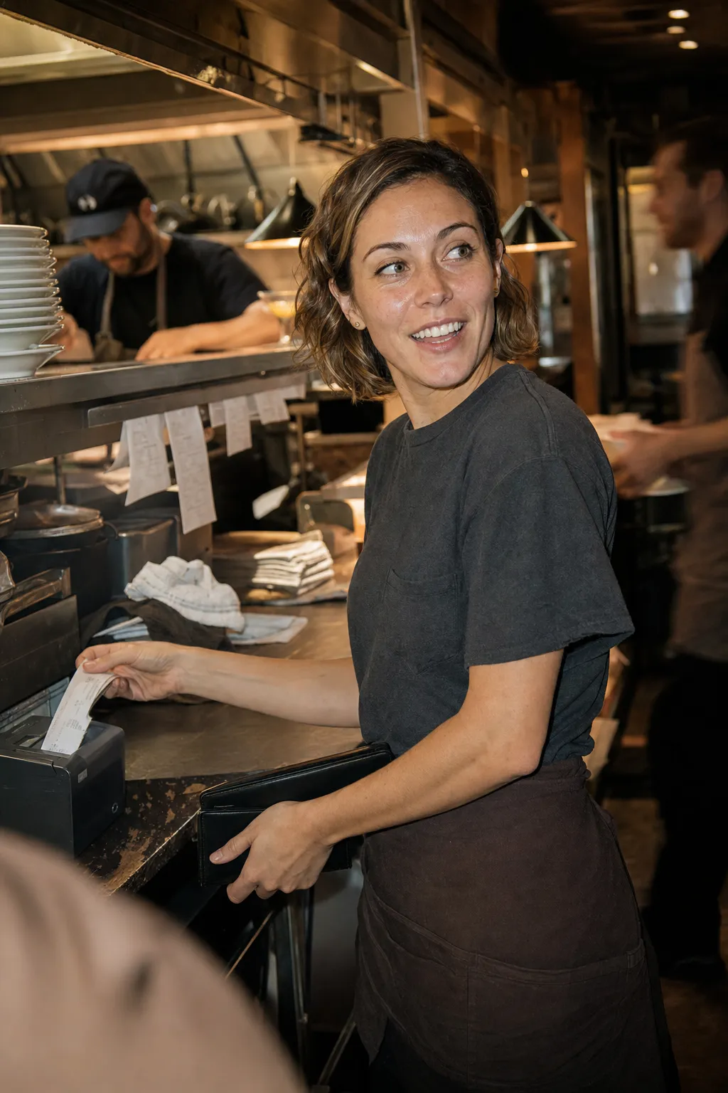 Smiling server in a dark t-shirt and apron collecting a printed receipt at a busy restaurant kitchen pass.