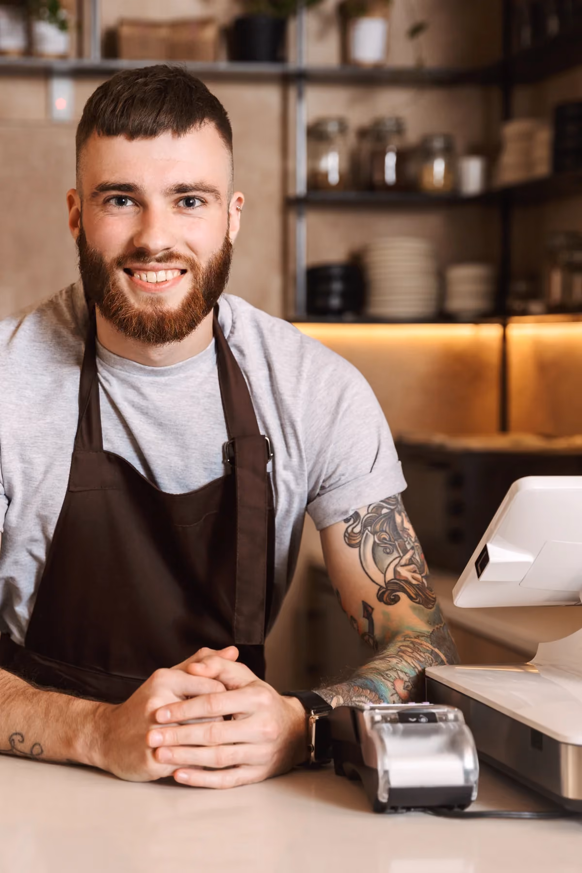 Smiling barista in an apron leaning on a café counter beside a POS terminal and card reader.