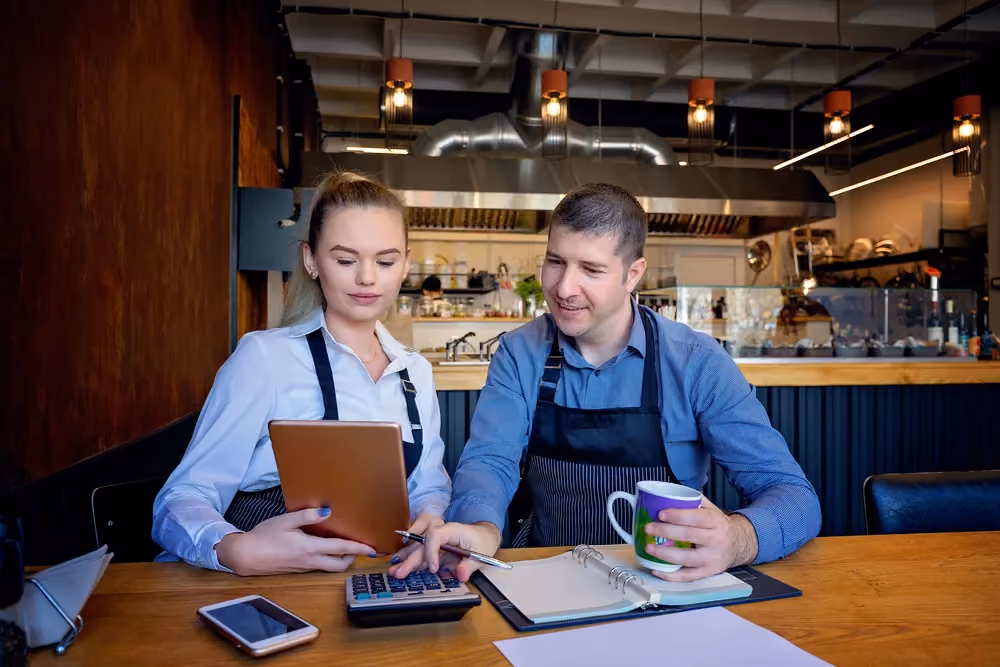 Two restaurant staff working out tip splits on a tablet and laptop.