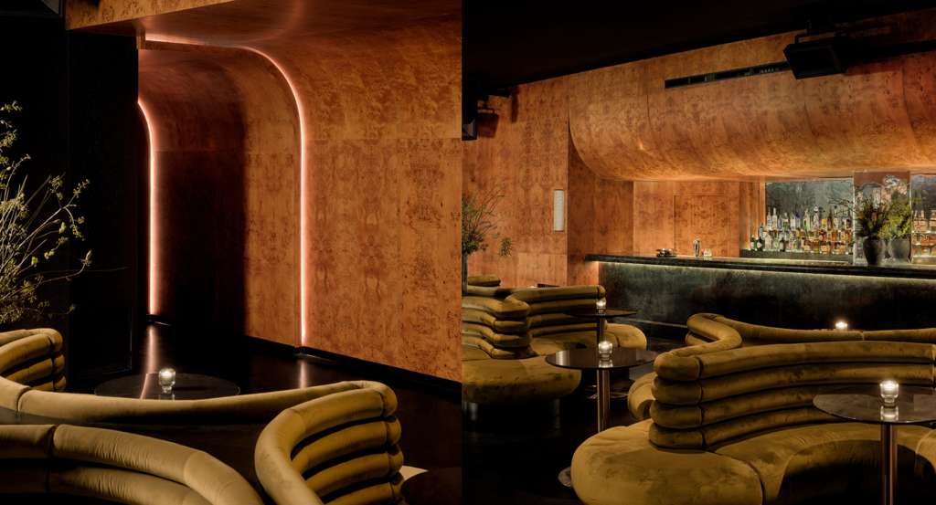 Modern lounge with curved olive-green velvet sofas, round black tables, and softly lit wooden-paneled walls surrounding a bar with bottles and floral decor.