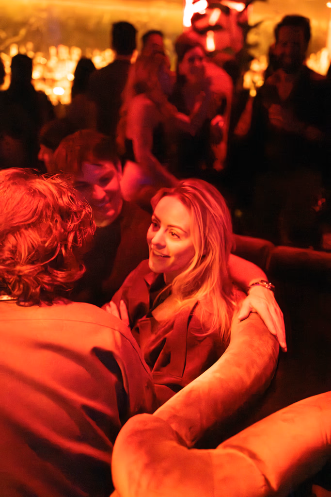 Three people sitting closely on a couch in a dimly lit lounge with warm red and orange lighting, engaged in conversation.