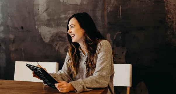 Smiling woman with long hair holding a tablet and sitting at a wooden table against a dark textured wall.