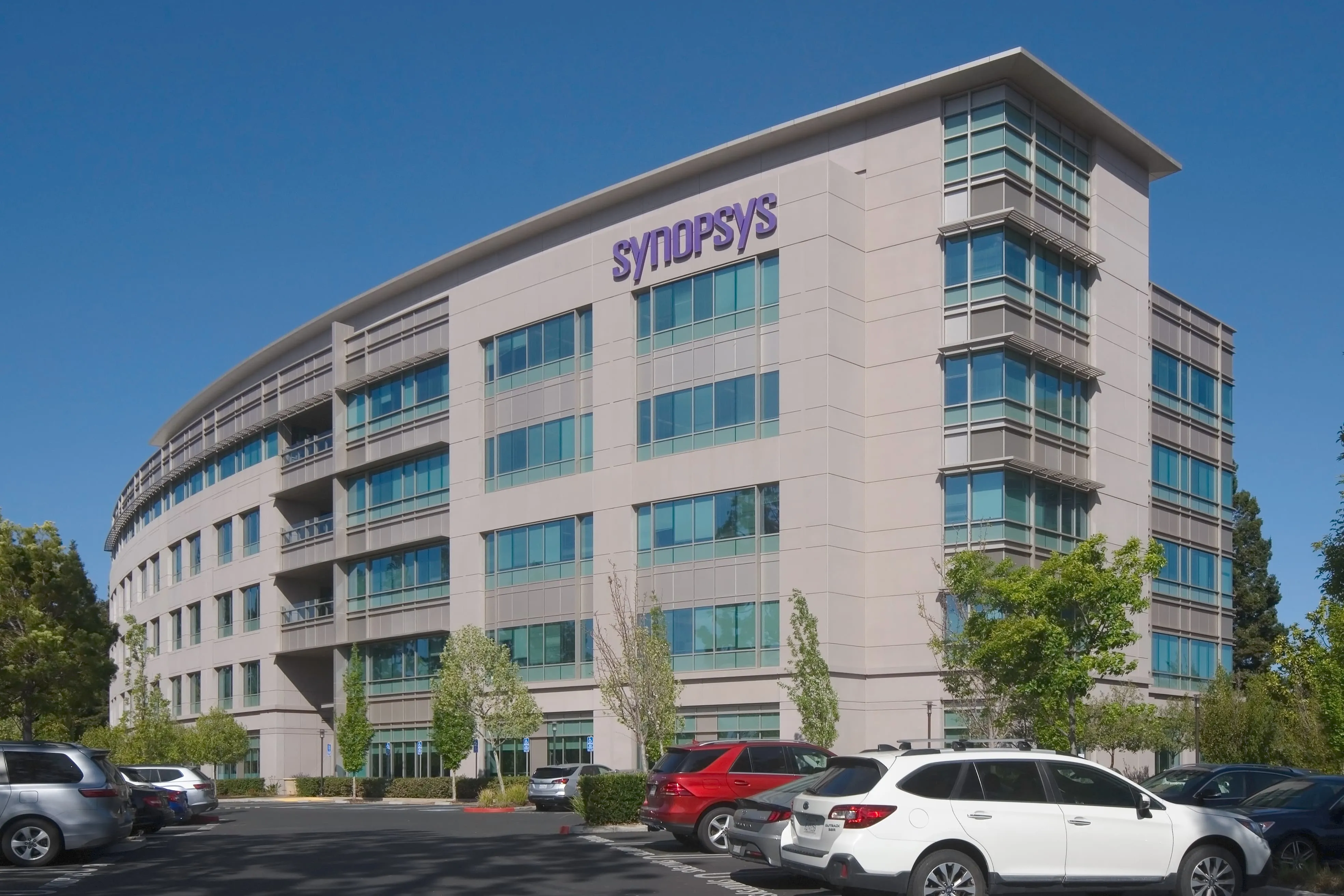 Modern multi-story Synopsys office building with many windows and a parking lot with cars in front under a clear blue sky.