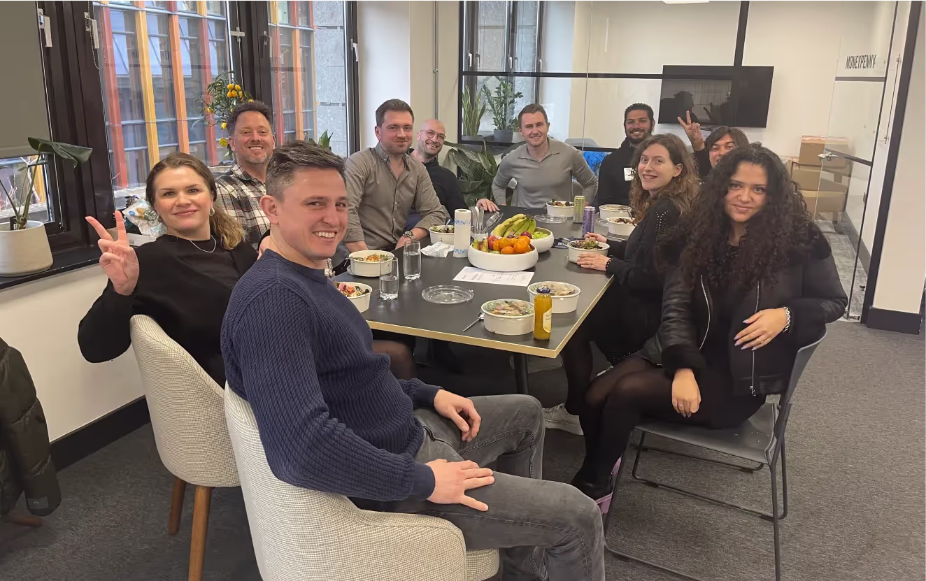 Group of ten people smiling and sitting around a table with food and drinks in a modern office setting.