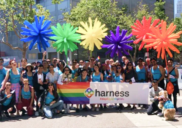 Group of people in blue shirts holding a rainbow flag and colorful star-shaped balloons at an outdoor event with trees and buildings in the background.