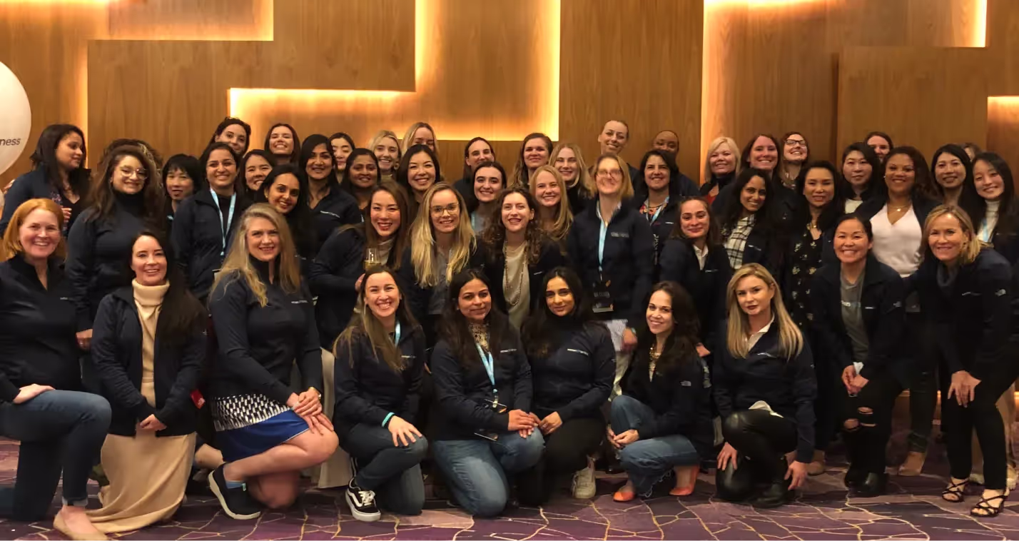 Large diverse group of women posing together indoors, many wearing matching dark jackets.