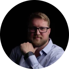Man with glasses and beard wearing a light blue shirt, resting his chin on his fist against a black background.