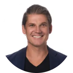 Smiling man with short brown hair wearing a dark blazer and black shirt against a white background.