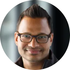 Smiling man with short dark hair and glasses, blurred background.