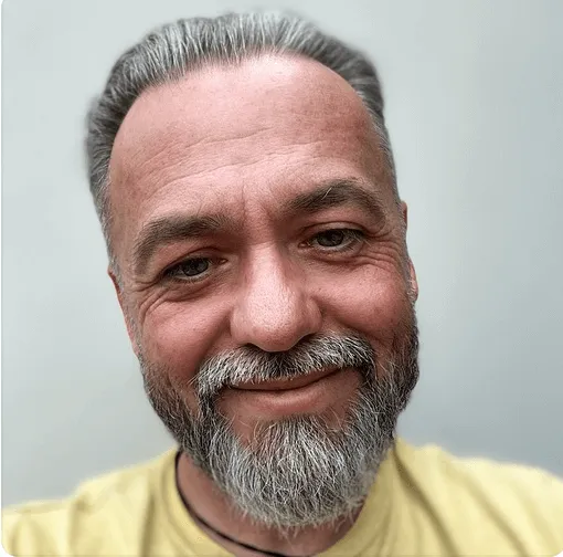 Smiling middle-aged man with gray hair and beard wearing a yellow shirt against a plain background.