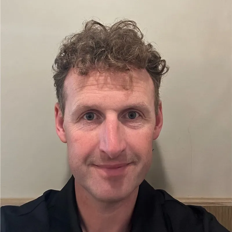 Man with curly brown hair and blue eyes wearing a black shirt, smiling in front of a beige wall.