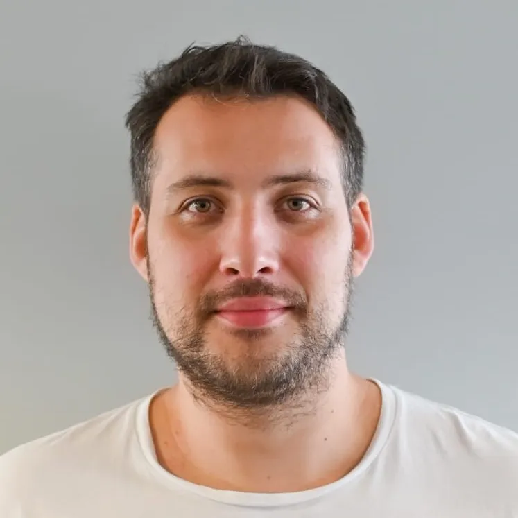 Portrait of a man with short dark hair, light beard, and wearing a white t-shirt against a neutral gray background.