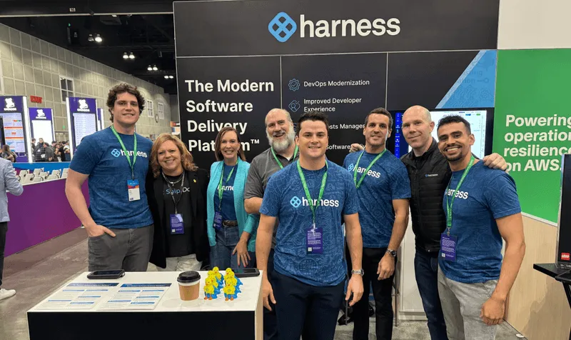 Group of eight people posing and smiling at a trade show booth with Harness logo and software delivery platform information.