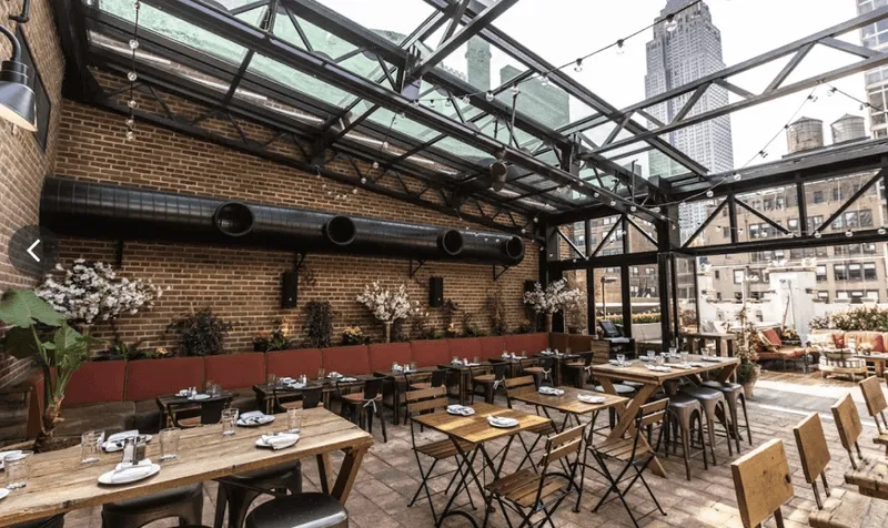 Urban outdoor restaurant patio with wooden tables and chairs under a glass roof, brick walls, potted plants, and city buildings in the background.