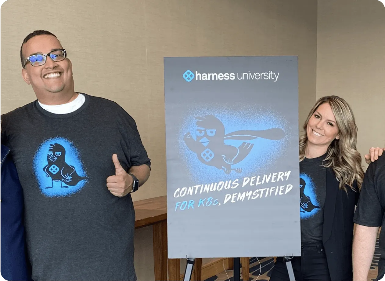 Two smiling people wearing dark t-shirts with a bird logo standing beside a poster that reads 'harness university CONTINUOUS DELIVERY FOR K8s, DEMYSTIFIED'.