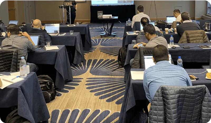 People seated at tables with laptops and notebooks attending a presentation in a conference room.