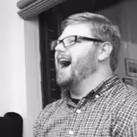 Man with glasses and beard laughing while wearing a checkered shirt indoors.