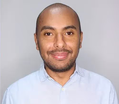 Headshot of a smiling man with a shaved head and short beard wearing a light blue collared shirt against a gray background.