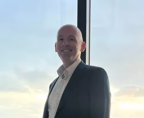 Smiling bald man wearing a suit jacket and white shirt standing indoors by a large window with a bright sky outside.