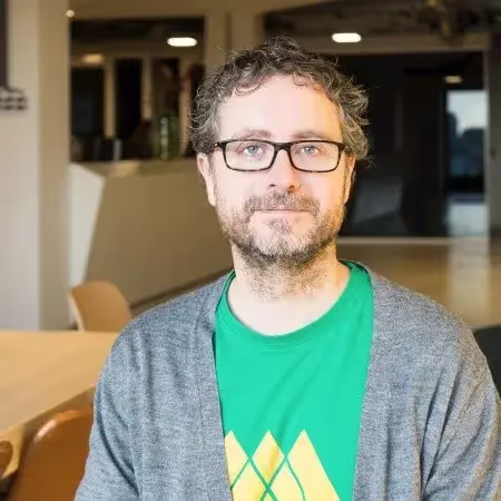Middle-aged man with glasses, curly hair, and a beard wearing a green shirt and gray cardigan in an indoor setting.