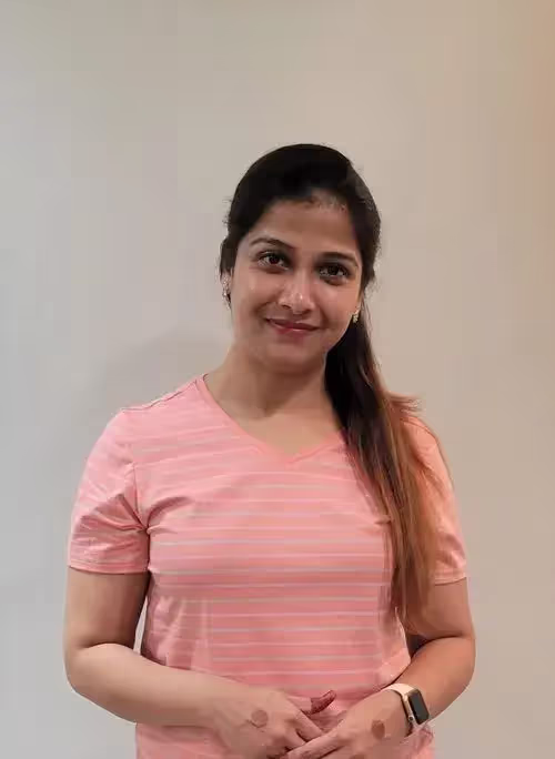 Woman with long hair wearing a pink striped t-shirt, smiling and standing against a plain light-colored wall.