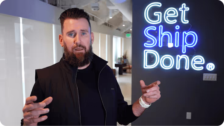Man with beard and black jacket in an office gesturing with hands next to a neon sign reading 'Get Ship Done.'