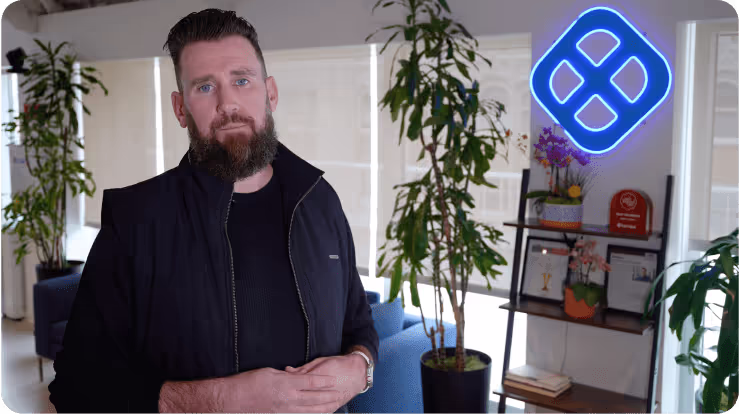 Man with beard and dark jacket standing in a modern room with plants, blue armchairs, and a glowing blue logo on the wall.