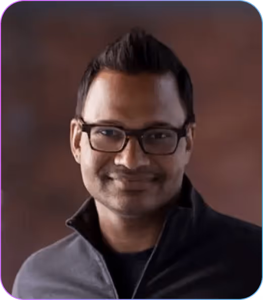 Portrait of a man with short dark hair, wearing glasses and a dark jacket, smiling against a blurred brown background.