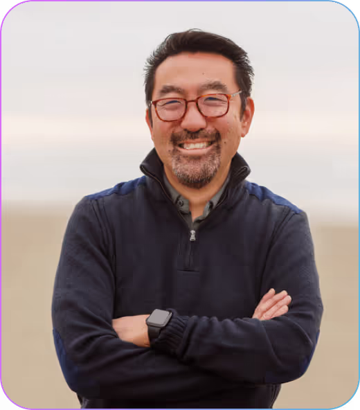 Smiling man with glasses wearing a dark zip-up sweater and a smartwatch, standing with arms crossed against a blurred neutral background.
