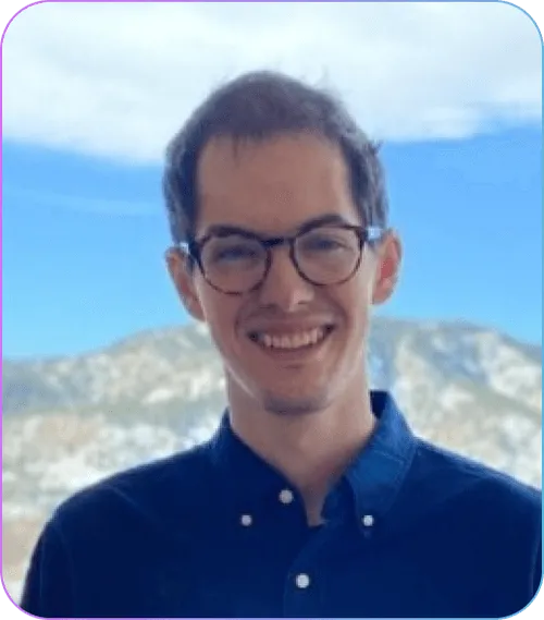 Smiling man with glasses and dark hair wearing a dark blue button-up shirt with a mountain landscape in the background.