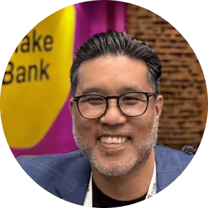 Smiling man with glasses and short black hair in front of a colorful background with a purple and yellow design.
