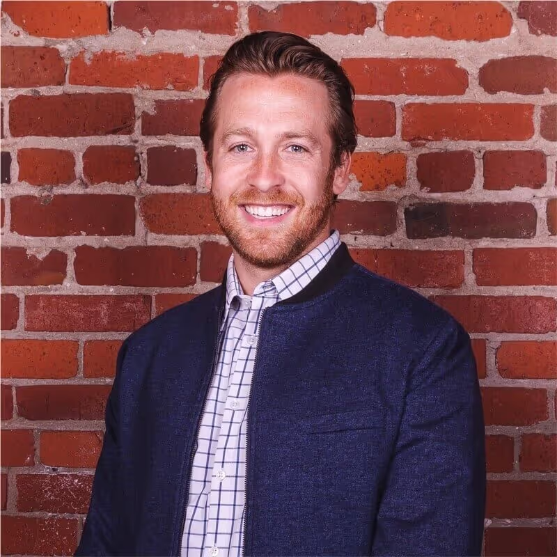 Smiling man with light brown hair and beard, wearing a navy jacket over a white checkered shirt, standing against a red brick wall.