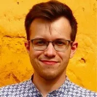 Young man with glasses and short dark hair smiling in front of a textured yellow wall.