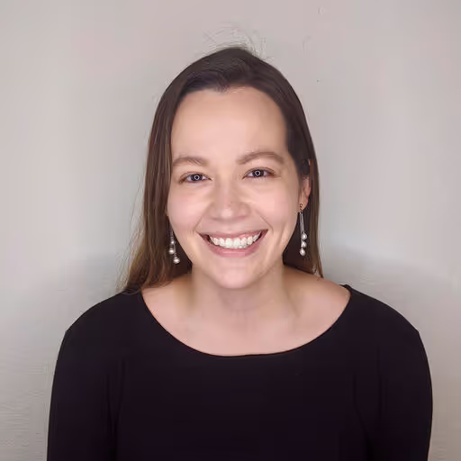 Smiling woman with long brown hair wearing pearl drop earrings and a black top against a plain background.