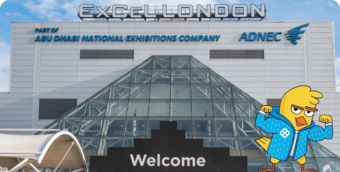 Exterior of ExCeL London exhibition center with glass pyramid entrance and a digital welcome sign, featuring a cartoon yellow bird in a blue jacket flexing its arms.