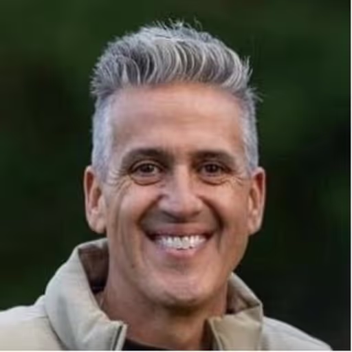 Smiling man with short gray hair wearing a light-colored jacket against a dark green background.