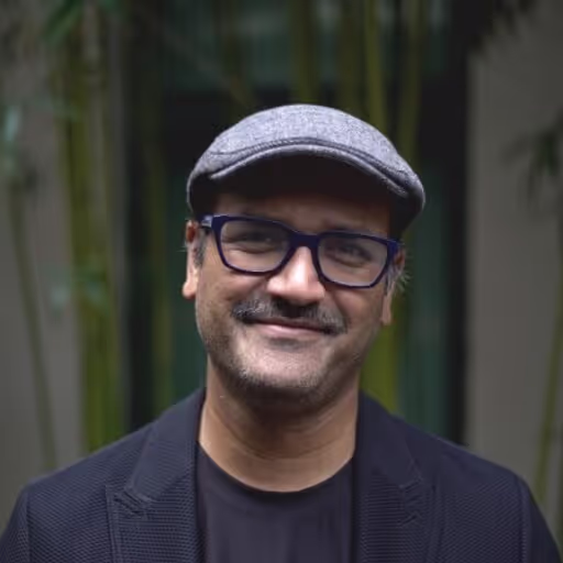 Smiling man wearing a gray flat cap, black glasses, and a dark blazer with a black shirt, standing outdoors with blurred green plants in the background.