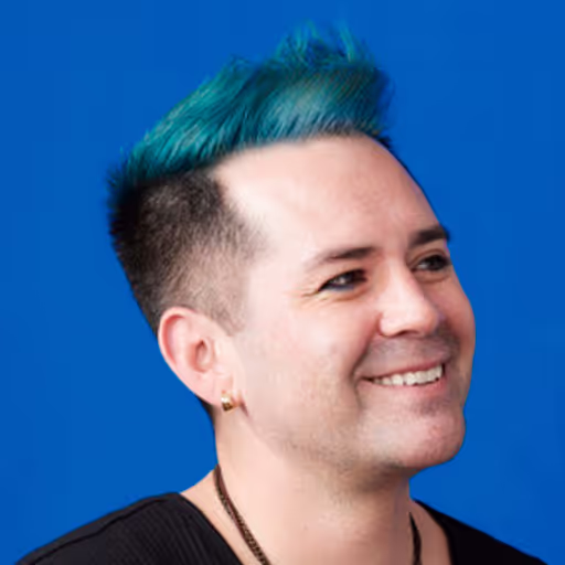 Smiling man with teal and black hair against a solid blue background.