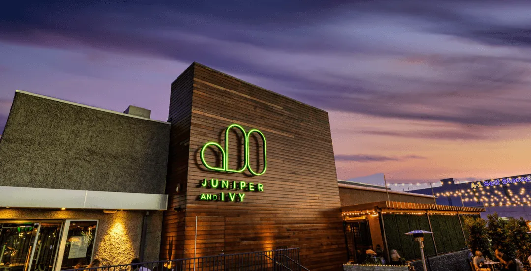 Exterior of Juniper and Ivy restaurant with a glowing green sign at dusk and people dining outside under string lights.