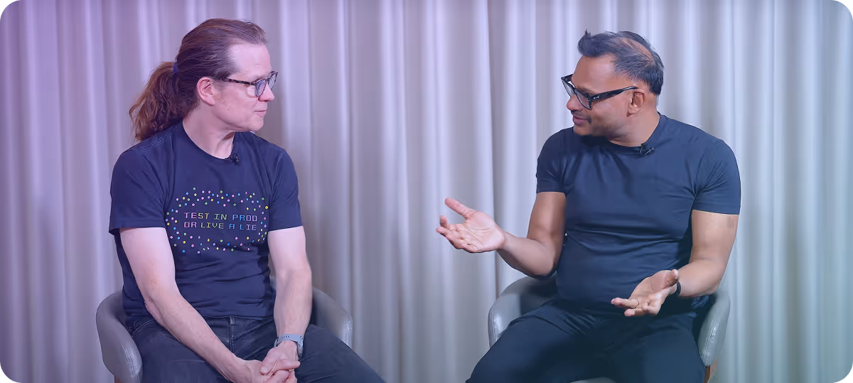 Two men seated and engaged in a conversation against a backdrop of light-colored curtains, one with glasses and long hair in a ponytail wearing a black t-shirt, the other with short hair and glasses wearing a dark t-shirt.