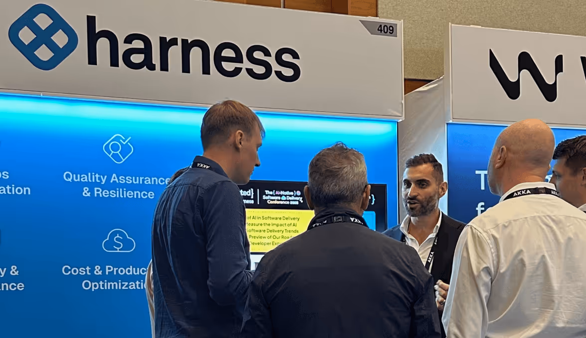 Four men standing and conversing at the Harness booth in an exhibition or conference setting.