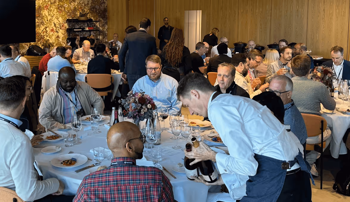 People seated at round tables in a dining room, with a waiter serving wine bottles.