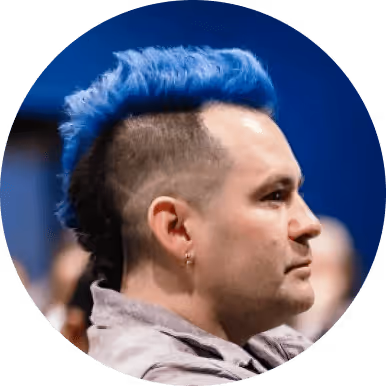 Profile of a man with vibrant blue mohawk hairstyle and an earring, wearing a light-colored shirt.